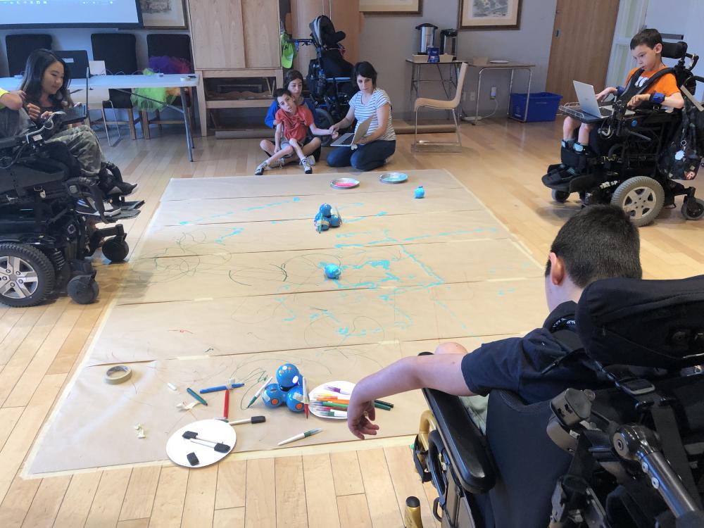 Students on mobility devices gathered around a floor level activity, trying to ove robots with programs they have built in Weavly. Students on mobility devices using their laptops to create a program to move robots on the floor to draw geommetric shapes. etric